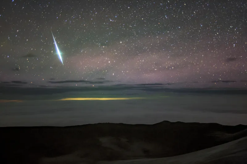 Une boule de feu blanc-bleu traverse un ciel étoilé rosé, au-dessus de nuages éclairés de jaune et d’une crête sombre
