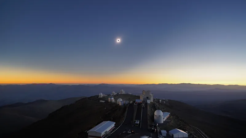 Petit disque noir cerclé de lumière dans un ciel de crépuscule bleu et orange, au-dessus d’un observatoire