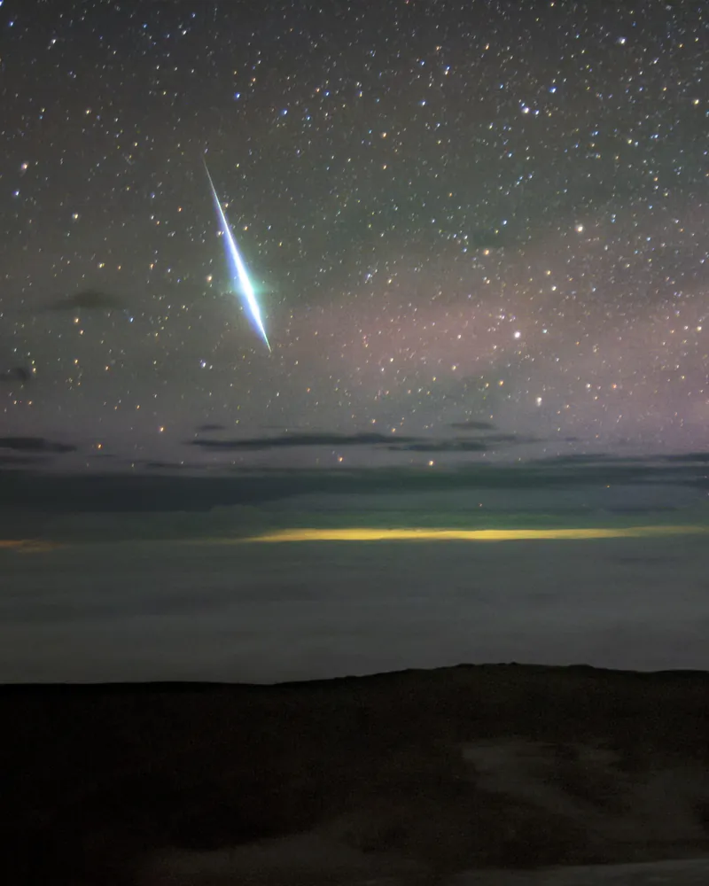 Une boule de feu blanc-bleu traverse un ciel étoilé rosé, au-dessus de nuages éclairés de jaune et d’une crête sombre