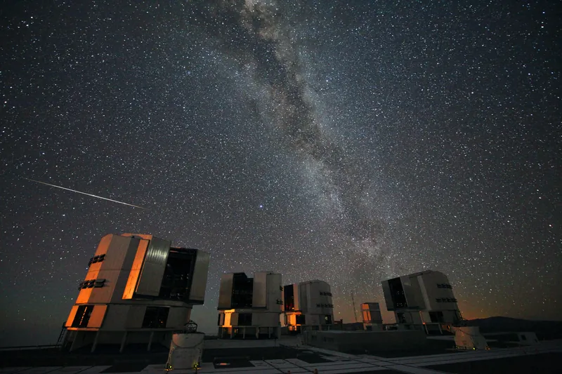 Un long météore fin raye le ciel à gauche de la Voie lactée, au-dessus de quatre coupoles blanches du VLT éclairées d’orange