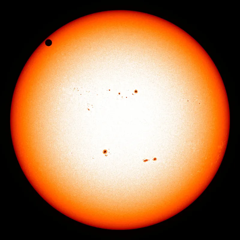 Le disque orange du Soleil, parsemé de taches, et la petite silhouette noire de Vénus près de son bord, en haut à gauche