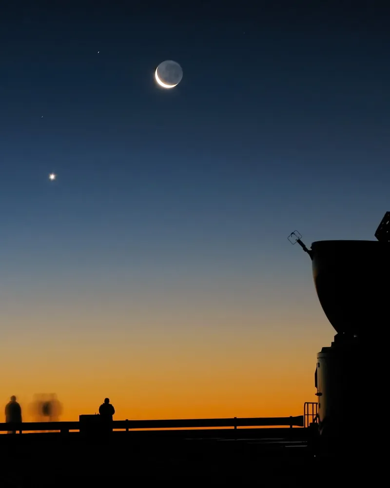 Un fin croissant de Lune et, à sa gauche, Vénus, point blanc, dans le ciel du couchant ; à droite, un télescope en silhouette