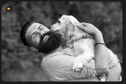 Homme souriant enlaçant son grand chien blanc dans un jardin, photo en noir et blanc